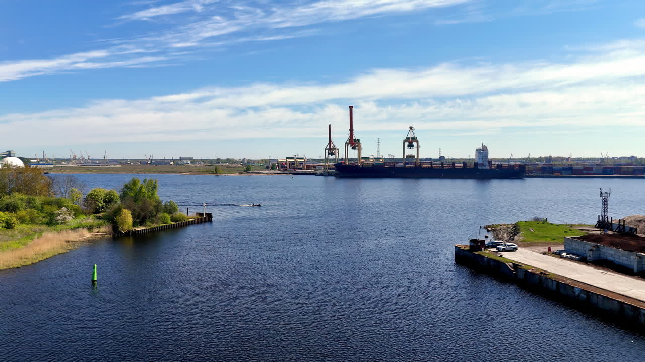 A calm harbor channel leads toward a container terminal where cranes service a vessel, with shoreline piers and a green marker framing the waterway in a steady wide drone view