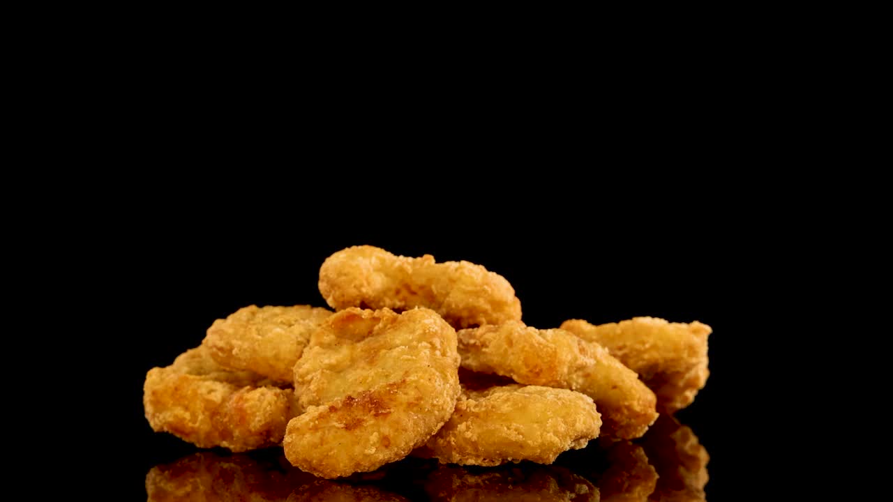 Multiple hands reach in to pick up golden, crumbed chicken nuggets from a stacked pile on a glossy black surface, under bright studio lighting