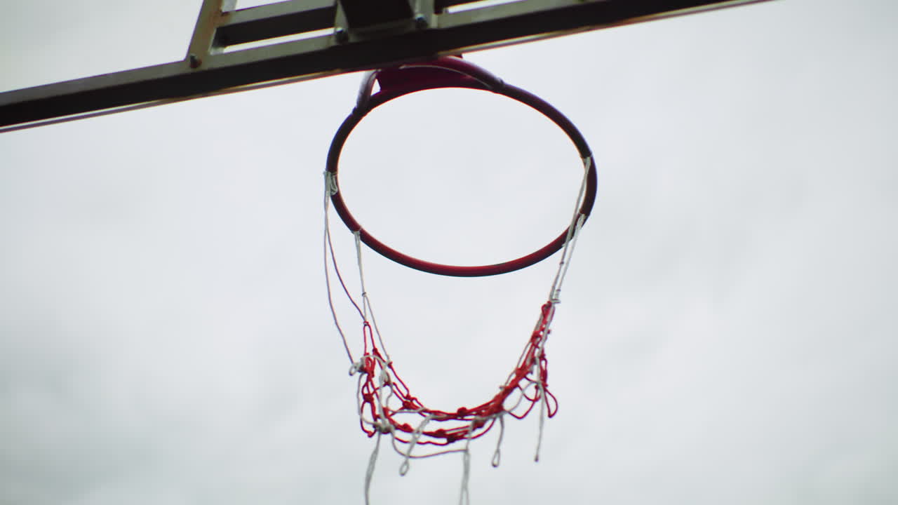Canestro da basket sopra la testa, il pallone sfreccia attraverso la rete sfilacciata, il canestro sobbalza, gocce di pioggia che accentuano il campetto urbano grintoso, primo piano del tabellone e della catena, movimento energico e trionfante
