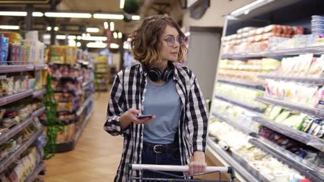 Shopping,-technology,-sale,-consumerism-and-people-concept---woman-with-smartphone-and-headphones-on-neck-walking-with-trolley-at-supermarket-or-store,-looking-at-mobiles-screen.-Front-view