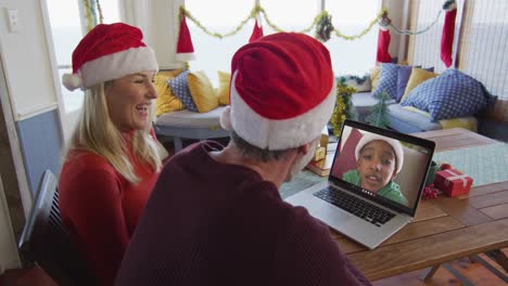 Smiling-caucasian-couple-with-santa-hats-using-laptop-for-christmas-video-call-with-boy-on-screen