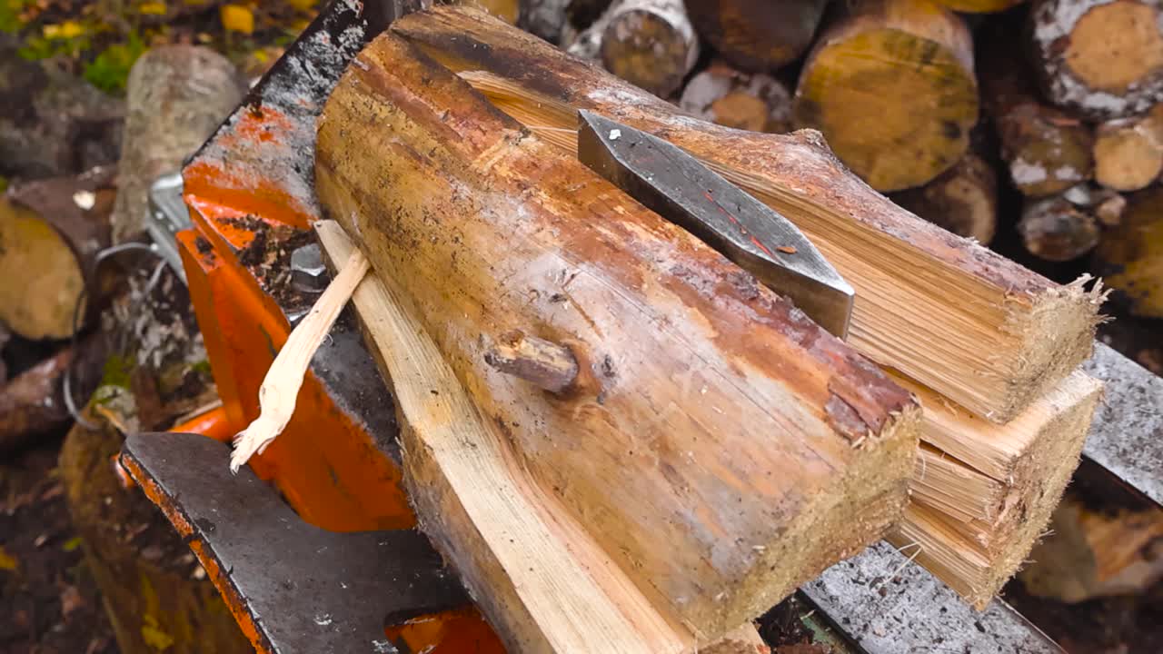 Close up view of a cut timber wooden pine log being split in pieces by a firewood splitting machine outdoors while cut wooden logs are in the vokeh blurry background at day time.Sawdust flying around