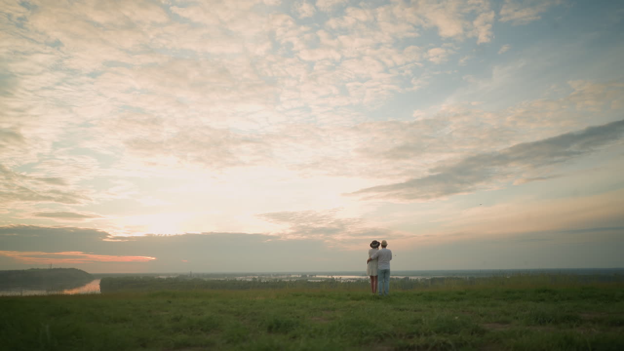 해가 지는 동안 호수 의 잔디 언덕에 서 있는 한 부부의 뒷면. 색 셔츠, 모자, 청바지를 입은 남자가 여자를 허리에 붙잡고 있다. 그녀는 검은 모자와 색 드레스를 입고 있다.