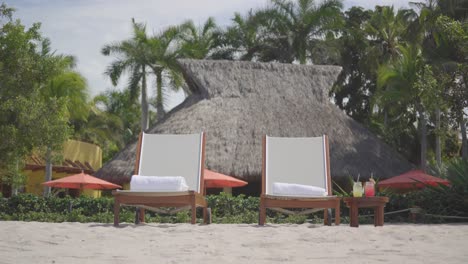 static shot of two lounge chairs on a tropical beach, two drinks on the side and a villa in the background