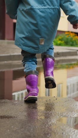 happy child runs across puddle on street closeup. healthy kid in jacket and waterproof boots plays outdoors on rainy day. active games after rainfall