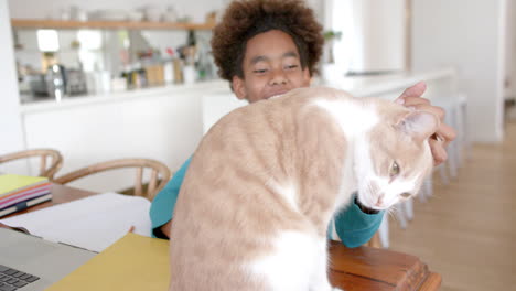 happy african american boy sitting at table, petting his pet cat, slow motion