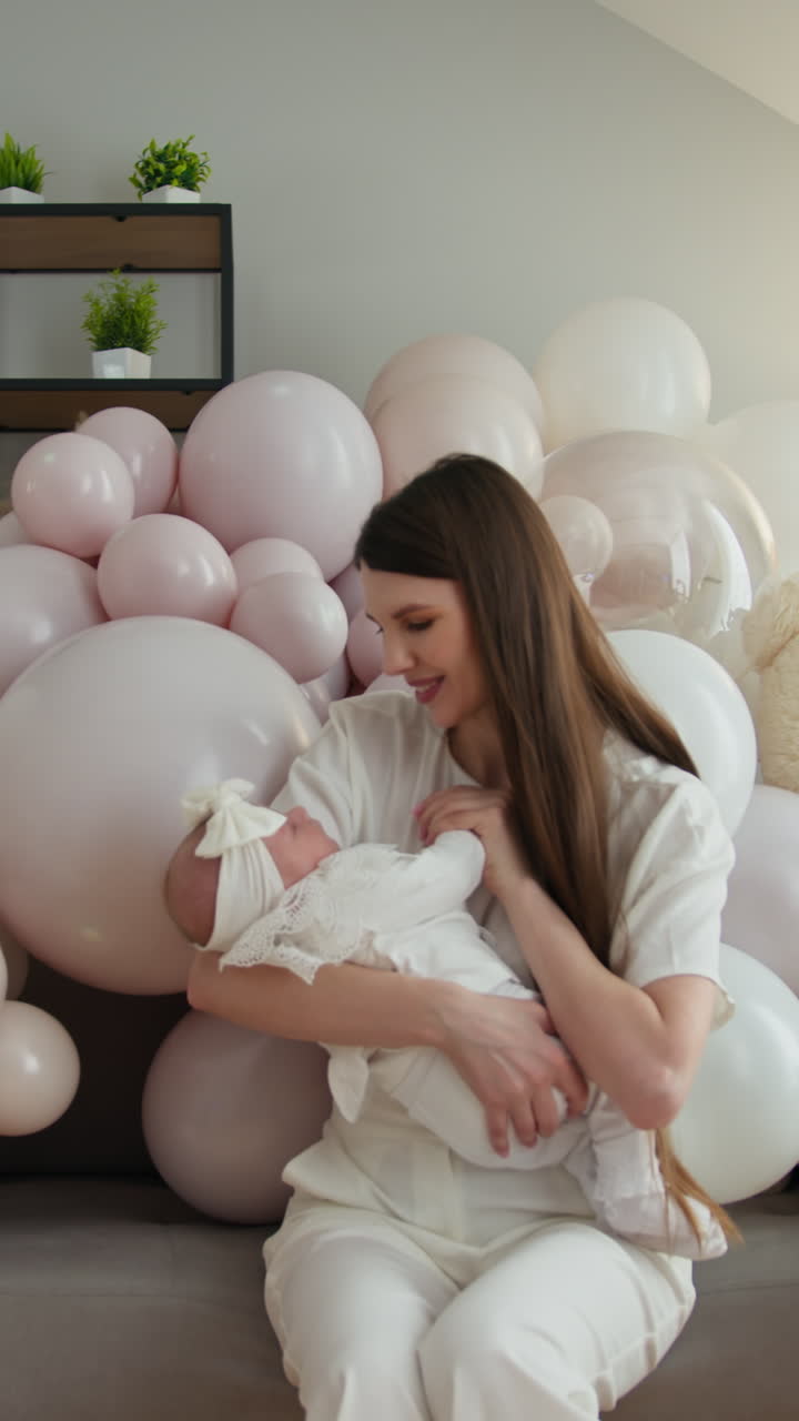 Madre culla amorevolmente la sua neonata, circondata da palloncini pastello, celebrando la nuova vita e