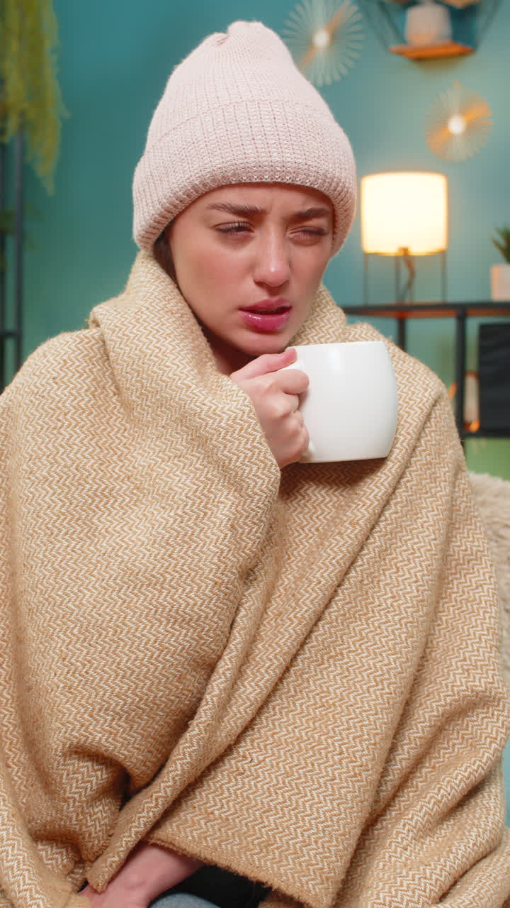 Young woman shivering on sofa under blanket wearing hat sipping hot tea hoping for gentle recovery