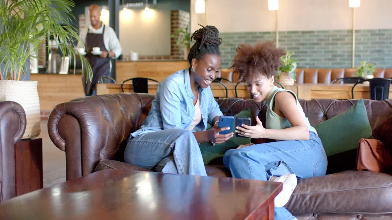 African American women sharing content, leaning, scrolling on sofa in cafe while staff passing tray