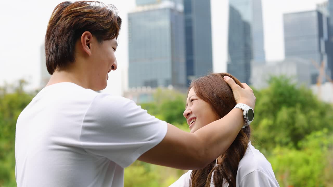 A young man gently touches his girlfriend's hair while smiling in a sunny urban park