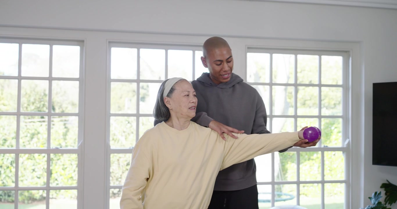 Caregiver placing purple dumbbell into Senior Asian woman, guiding side raises at home for rehab