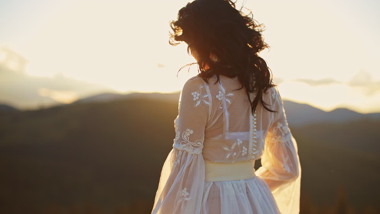 Long-haired lady wearing white dress standing high in the mountains. Lady is turning around at the backdrop of shining bright setting sun.