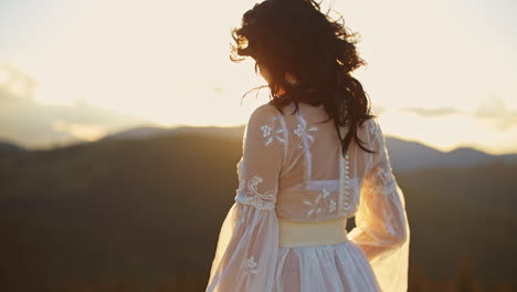 long-haired lady wearing white dress standing high in the mountains. lady is turning around at the backdrop of shining bright setting sun.