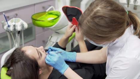 joven paciente de sexo femenino en el dentista con un espejo para comprobar los resultados del procedimiento. joven dentista de sexo femenino en guantes y máscara acercándose con herramientas.