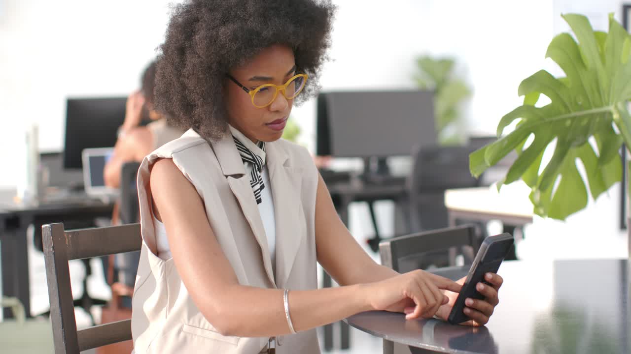 African American woman tapping smartphone while reading message at coworking table, smiling