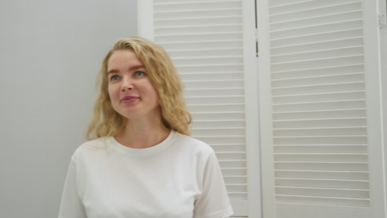 Confident client with long blonde hair enters white lit salon smiling warmly while interacting with beautician. Bright setting features white decoration and glowing signage in background near doorway