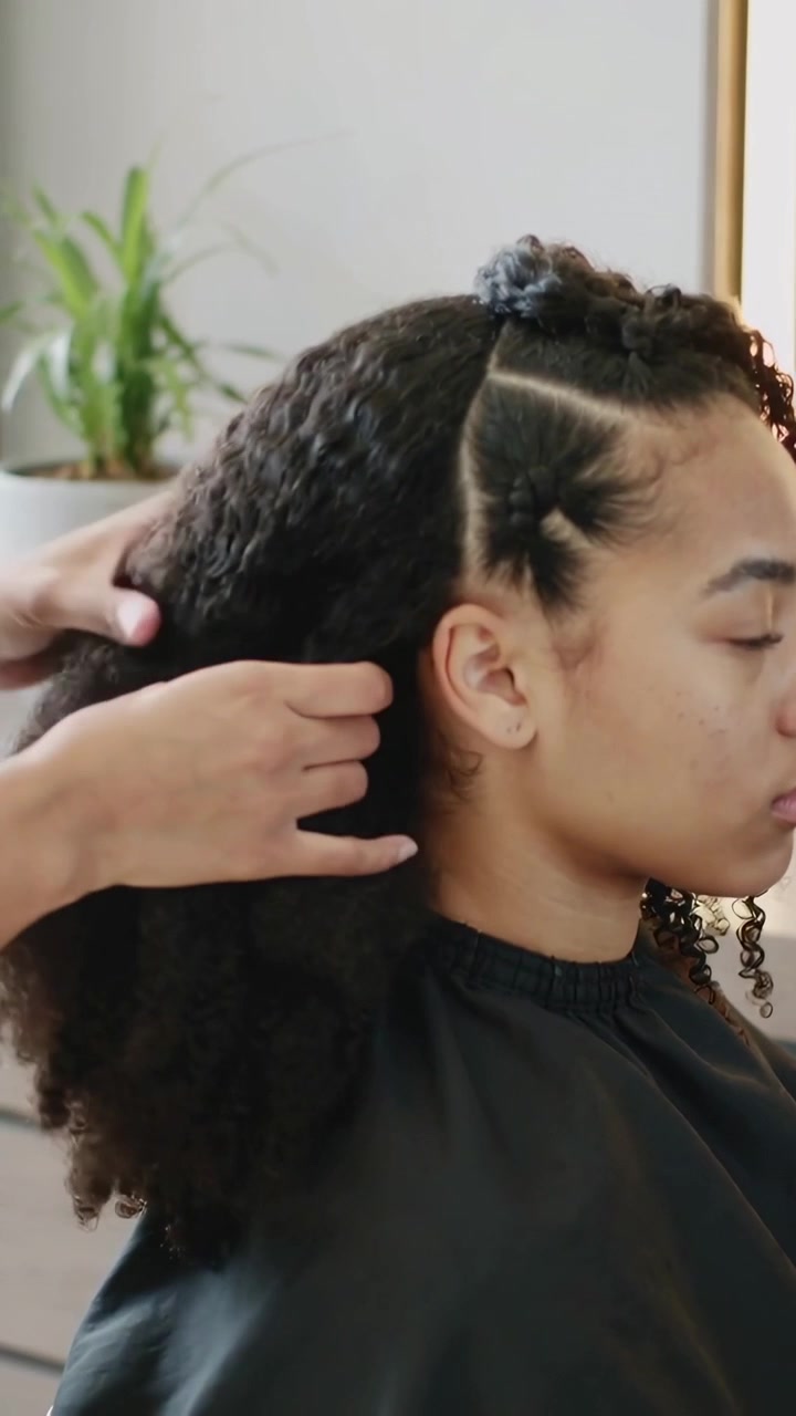 Video verticale: Stilista che solleva una parte e acconcia i capelli di una donna in un salone, creando una torsione con un mantello nero.