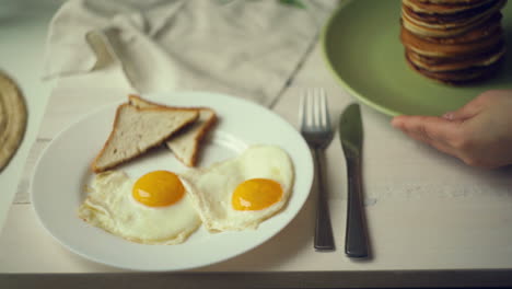 Woman-hands-put-plate-with-pancake-stack-on-table-near-plate-with-fried-eggs