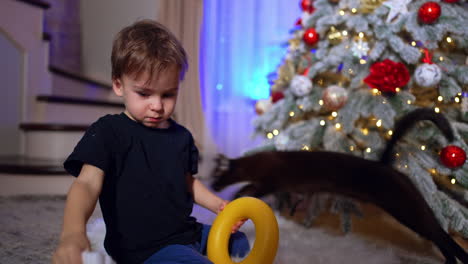 two year old toddler takes off santa cap. funny baby puts yellow toy ring to his face. christmas tree at backdrop.