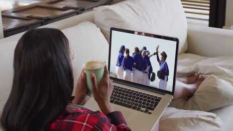 Compuesto-De-Mujer-Sentada-En-Casa-Sosteniendo-Un-Café-Viendo-Un-Partido-De-Béisbol-En-Una-Computadora-Portátil