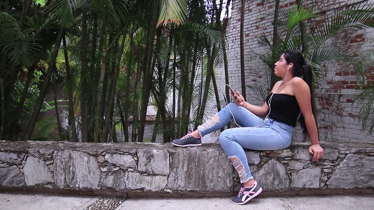niña sentada en un muro de piedra, escuchando música desde su teléfono, a través de un auricular con cable