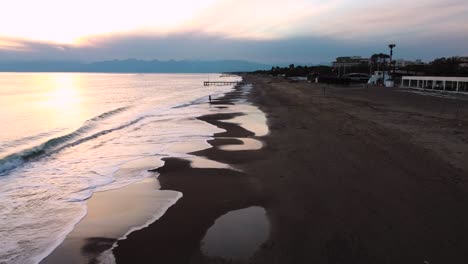 puesta de sol nublada sobre la playa, pequeñas olas y una chica parada frente al agua, tiro con drones