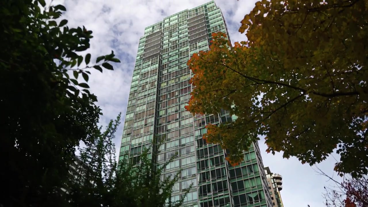 Paso rápido a través de árboles verdes y naranjas en primer plano para revelar un edificio de apartamentos alto que se alza solitario en la ciudad con ventanas de espejo azul claro en un día nublado en Vancouver