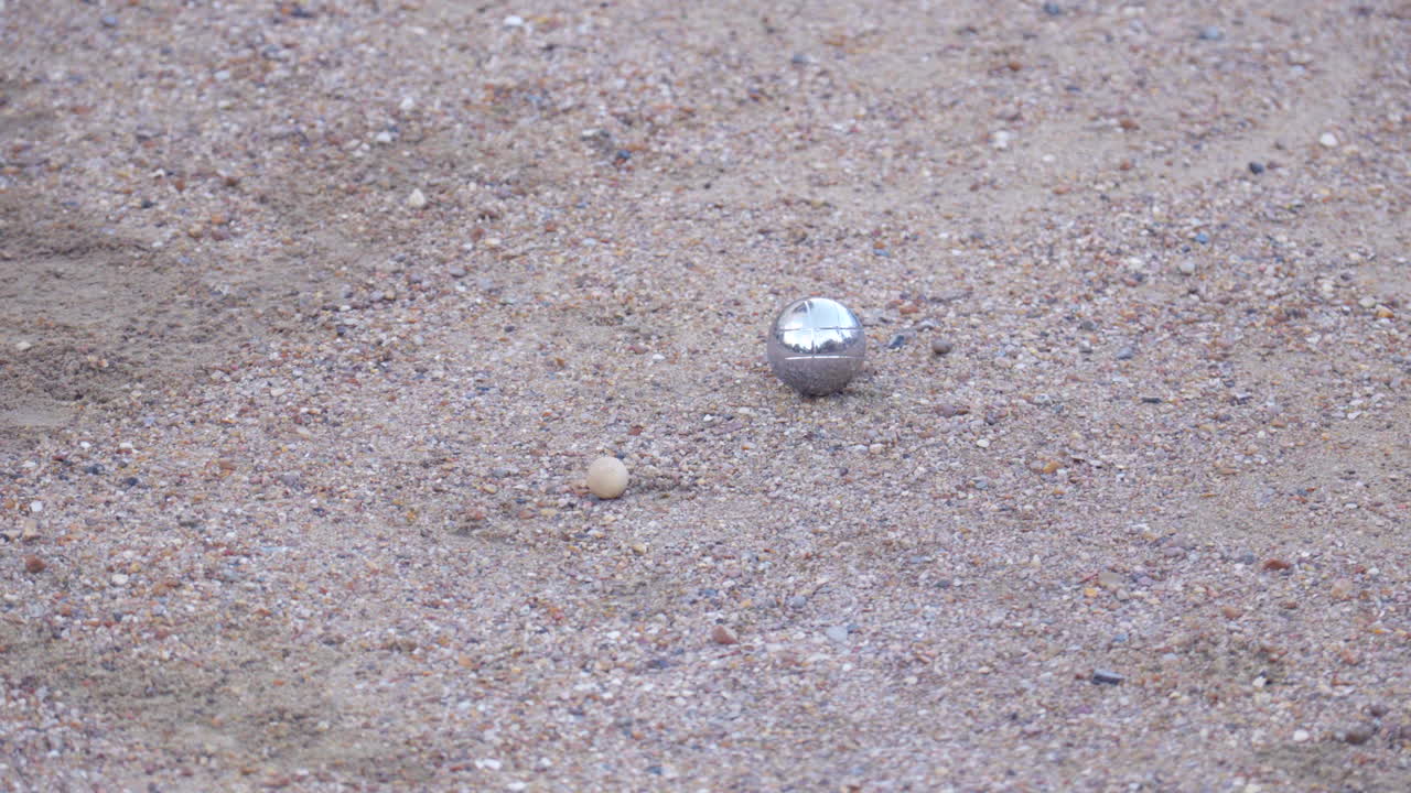pétanque ball rolls into frame, nudges the cochonnet, and continues its path out of the frame.