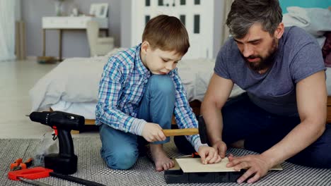 bearded father and young cute son making birdhouse of wooden sheets at home. childhood and parenthood concept