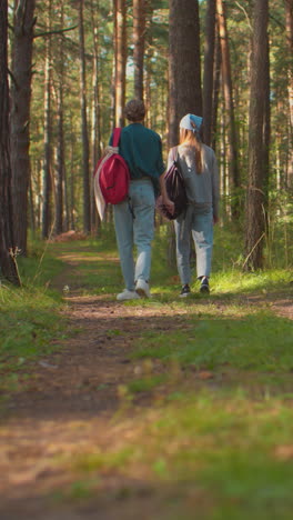 vista trasera de dos hermanas caminando por un sendero forestal rodeado de exuberante vegetación, una lleva una corbata de cabello azul, llevando una mochila negra, mientras que la otra tiene una mochila roja, árboles altos en el camino