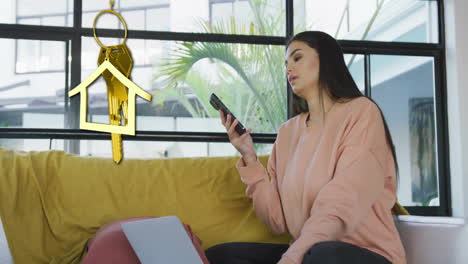 animation of gold house key and key fob over happy biracial woman using smartphone at home