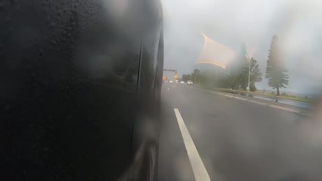 Rain-running-down-on-camera-as-a-black-car-drives-along-a-highway-during-a-rainy-late-afternoon-with-the-camera-facing-the-front-wheel-and-on-coming-traffic