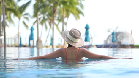 Bella-Mujer-Con-Sombrero-Para-El-Sol-Entrando-En-El-Agua-De-La-Piscina-En-Un-Resort-De-Isla-Tropical,-Portátil-En-Cámara-Lenta