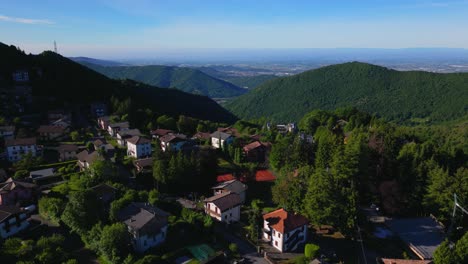 wide aerial view of a hillside village with traditional houses, forested slopes, and distant urban plains under a clear sky. shot at selvino, italy (selvino, italia)