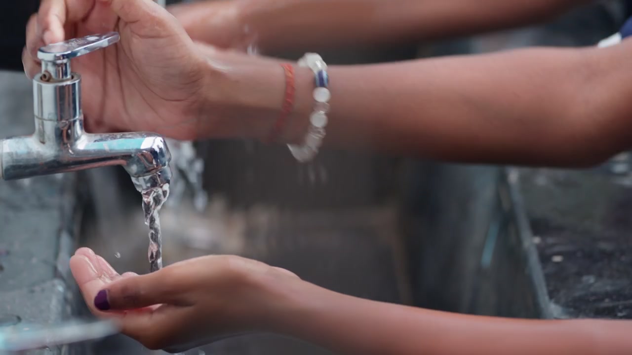 Diversi bambini si lavano le mani insieme sotto l'acqua corrente di un rubinetto cromato presso una stazione di lavaggio mani all'aperto. L'ambientazione è una scuola rurale in India con studenti che praticano l'igiene quotidiana.