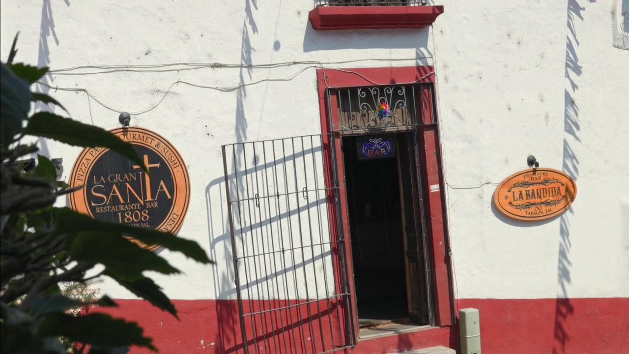 La luce del giorno rivela una classica facciata coloniale rossa e bianca con le insegne 'La Gran Santa' e 'La Bandidita', un ingresso in ferro battuto, ombre frondose e muri in stucco testurizzato. Nessuna persona visibile