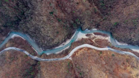 Carretera-Sinuosa-Que-Recorre-Un-Accidentado-Paisaje-Otoñal-Paralelo-A-Un-Río,-Vista-Aérea-De-Arriba-Hacia-Abajo