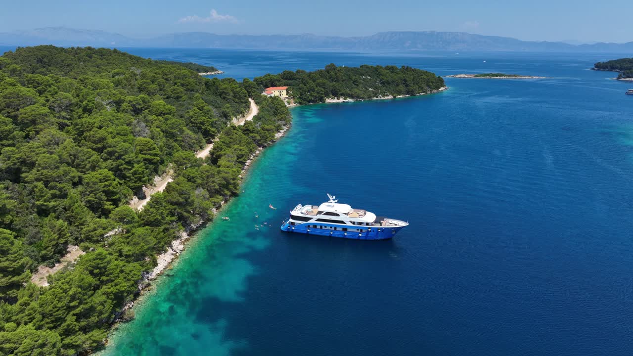Yacht di lusso ancorato in acque limpide e turchesi al largo della costa boscosa di Mljet, Croazia, con persone che nuotano vicino all'imbarcazione.