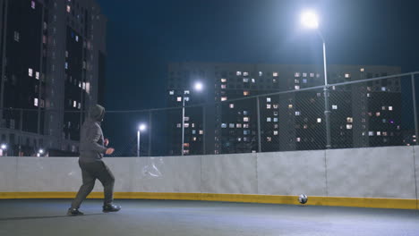vista trasera de un atleta con capucha jugando al fútbol contra la pared bajo la iluminación brillante del estadio por la noche, la pelota de fútbol cerca de la pared refleja la silueta, con el paisaje urbano y los edificios iluminados