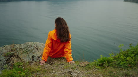 woman in orange jacket contemplating lake view from cliff