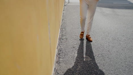 Man-in-beige-pants-and-brown-loafers-walking-along-a-sunlit-street-next-to-a-yellow-wall