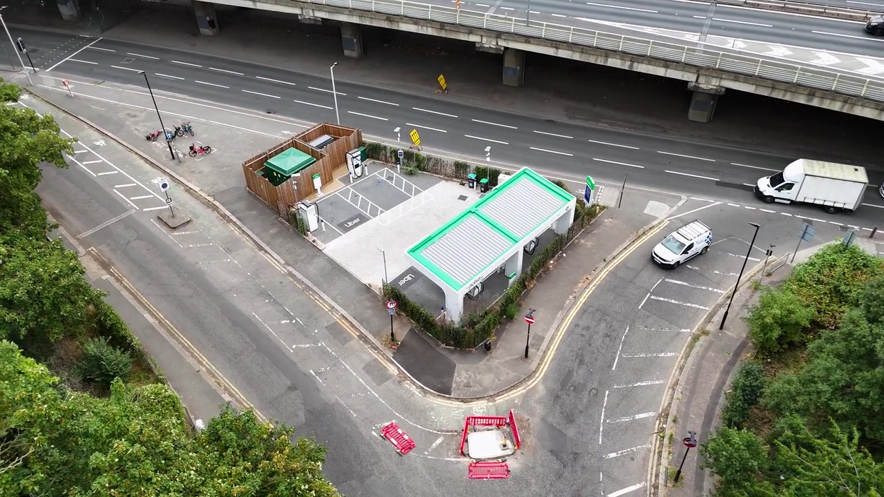Stazione di ricarica rapida per veicoli elettrici con tettoie a tetto verde, situata su una rampa di accesso sotto un'autostrada urbana.