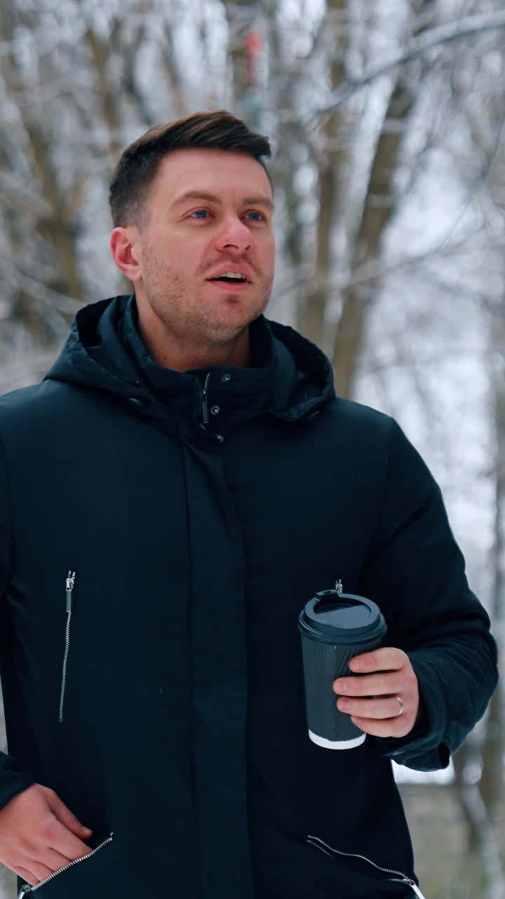 Man wearing warm black winter jacket walks outdoors carrying a paper cup. Man drinks coffee walking in winter park. Vertical video
