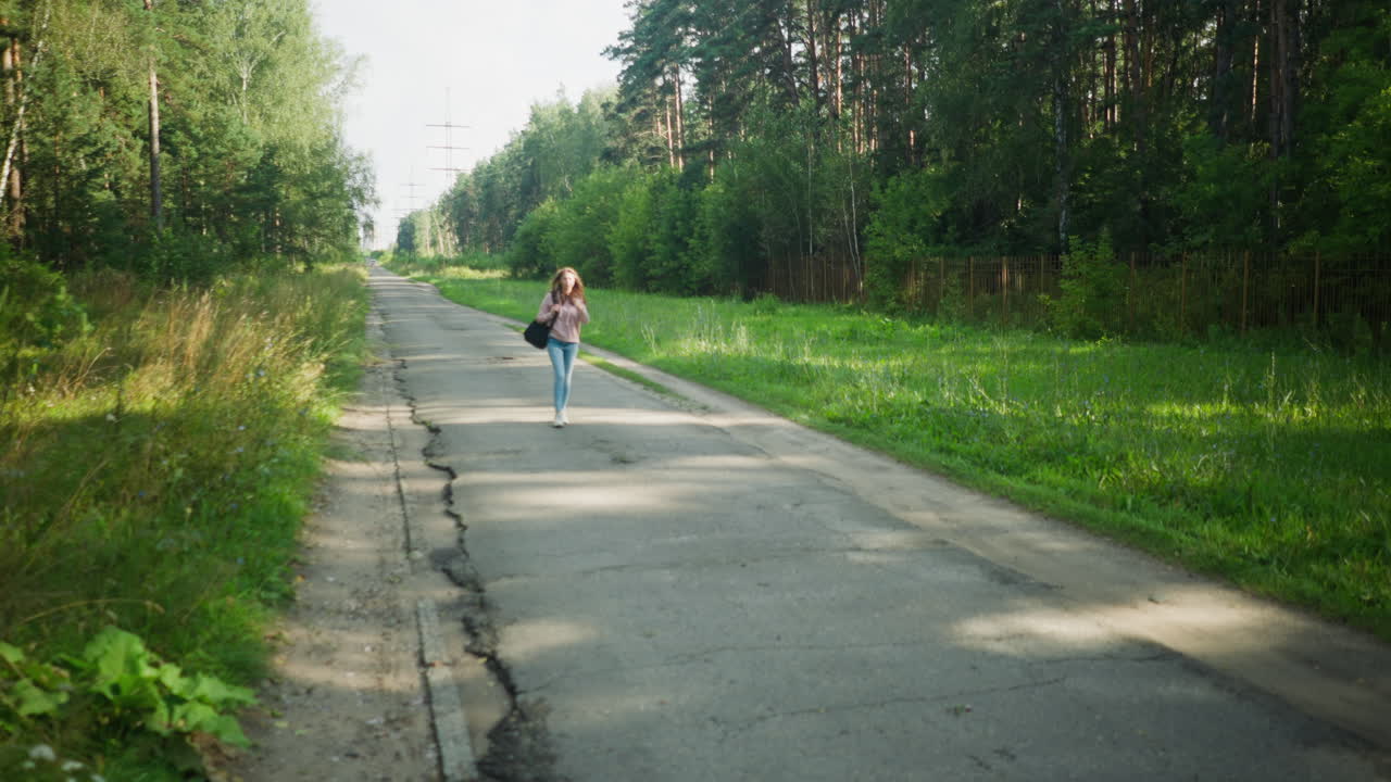 Young woman in pink hoodie and jeans walks alone along cracked rural road bordered by lush green grass and dense forest, carrying black shoulder bag and appearing calm and focused in daylight scene