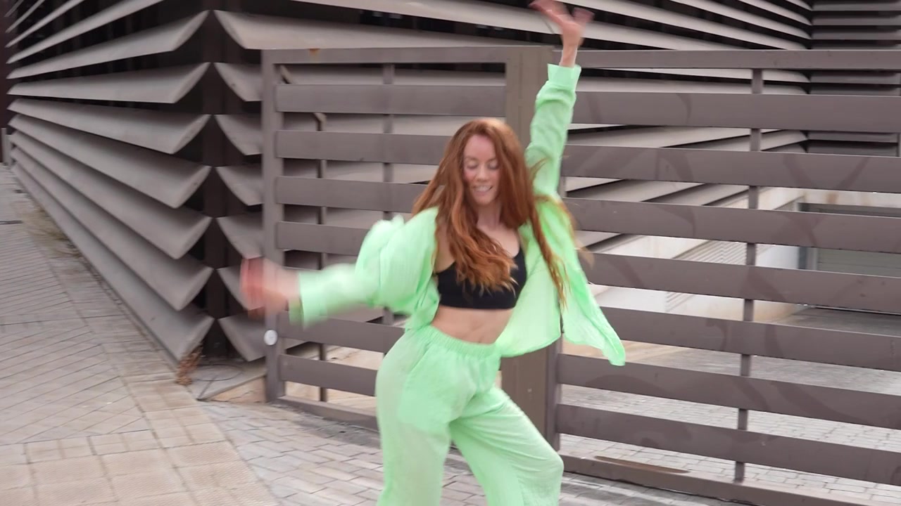 A lively woman dances energetically outside wearing a stylish green outfit with an urban background