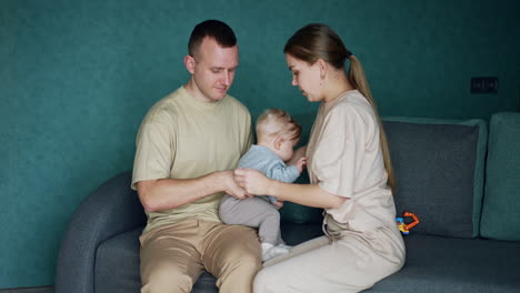 Woman puts on a sweater on the little infant baby sitting on dad's laps. Parents sitting on the sofa dress the cute blond child.
