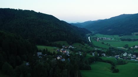 evening aerial drone footage of a small village surrounded by hills and forests, with a river flowing through the village