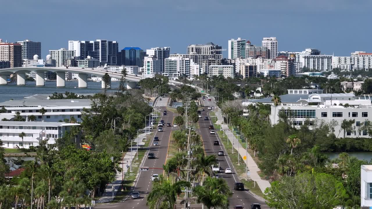 Aerial establishing shot of traffic scene on road with bridge and Sarasota Skyline in distance. Aerial backwards wide shot. Palm tree-lined highway following to St Armands Circle.