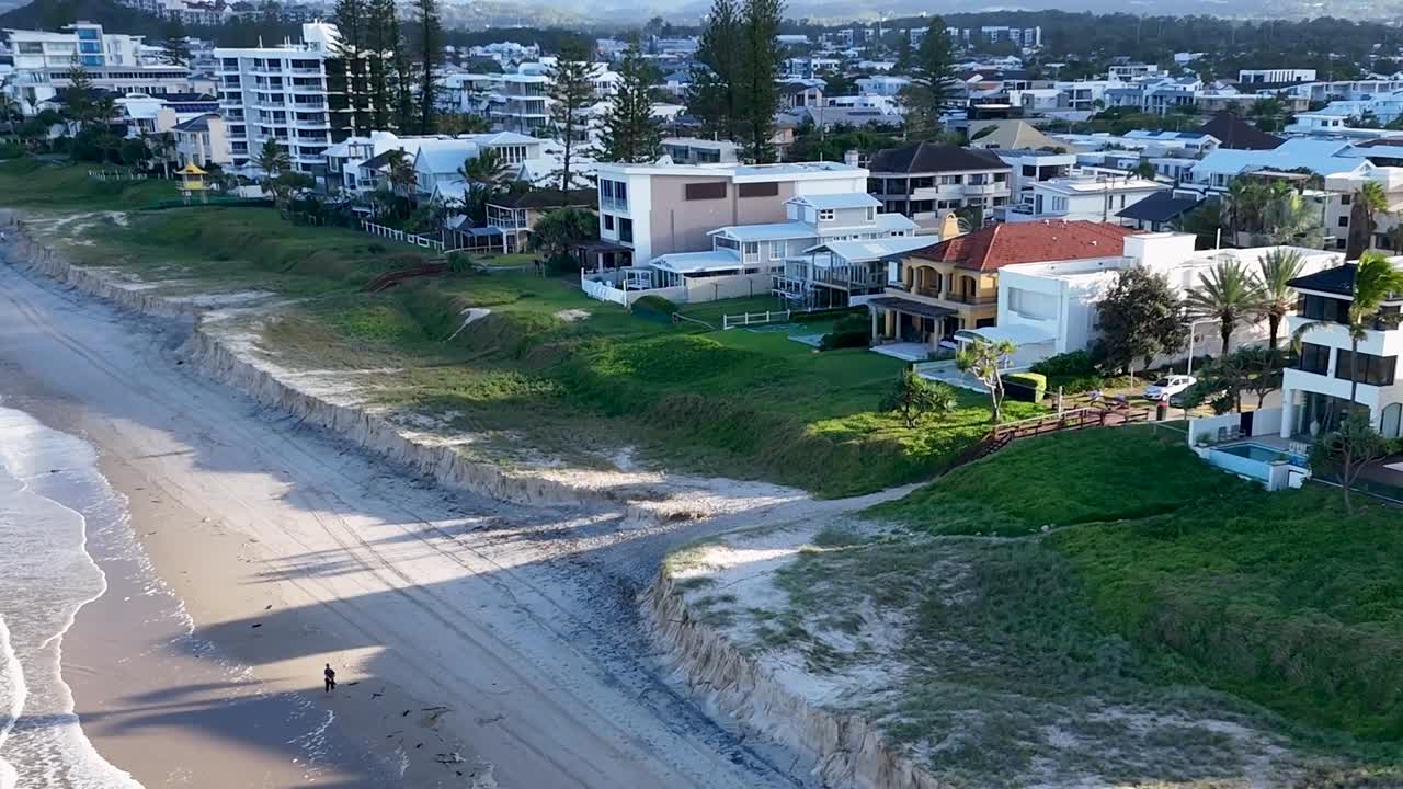 Imágenes de dron mostrando propiedades residenciales de lujo a lo largo de una playa de arena con olas suaves.
