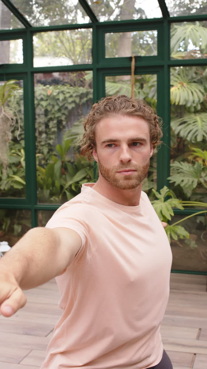 Vertical video: Practicing yoga, man stretching arm forward in glass house with greenery